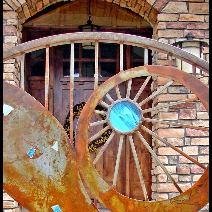A garden gate in weathered steel: one sweeping curve, a sunburst wheel and bright blue glass inlays.