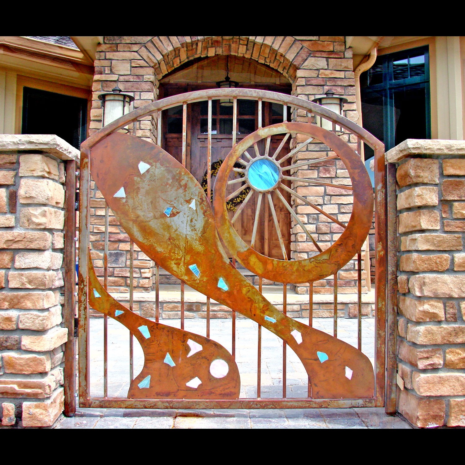 A garden gate in weathered steel: one sweeping curve, a sunburst wheel and bright blue glass inlays.